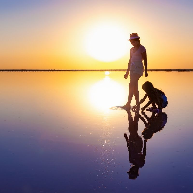 Two girls playing on the beach in shallow water with sunset behind them